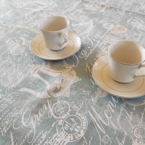 May include: Two white teacups with saucers on a light blue tablecloth with a white script pattern. The tablecloth has the words "Tea Time" and "Menu" printed on it.