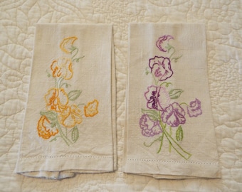 May include: Two embroidered linen tea towels with floral designs. One towel features yellow and orange flowers, while the other features purple and pink flowers.