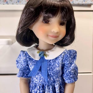 May include: A doll with dark hair and rosy cheeks wearing a blue floral dress with a white collar. The dress has short, puffed sleeves and a blue ribbon with a small flower accent. The doll's expression is gentle.
