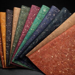 May include: A collection of textured paper sheets in various colors, including green, brown, and red, arranged in a fan shape. Each sheet has a unique pattern of gold-colored veins, creating a marbled effect. The background is black.