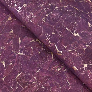 May include: Close-up of a roll of purple cork fabric with gold metallic accents. The cork has a mosaic-like pattern with irregular shapes and a textured surface. The gold leaf adds a touch of luxury to the design.
