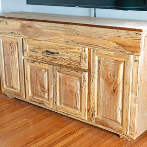 以下が含まれることがあります： 薄い茶色の仕上げのウッドキャビネット。4つのドアと黒い取っ手付きの引き出しが1つあります。キャビネットは硬材の床の上に置かれています。