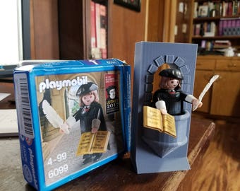 May include: Playmobil Luther figure and a 3D-printed tower. The Playmobil figure wears a black robe and hat, holding a quill and a book. The box features the Playmobil figure and the text "Luther 2017". The tower is gray and has a window.