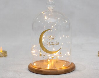 May include: A glass cloche with a wooden base, featuring a gold crescent moon design with the words "Eid Mubarak". Inside, delicate fairy lights illuminate the interior. The cloche is a decorative item, perfect for festive occasions.