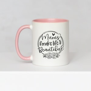 May include: White ceramic mug with a pink interior and handle. The mug features the text "Mums make life Beautiful" in a black, decorative font, surrounded by a floral wreath design. A heart shape is above the word "Mums".