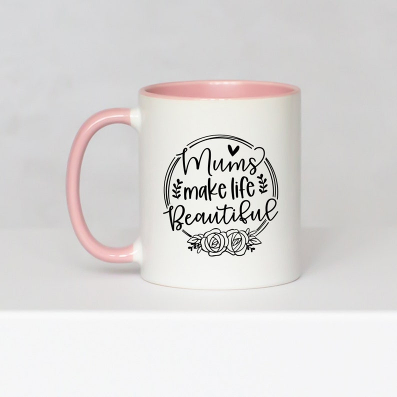 May include: White ceramic mug with a pink interior and handle. The mug features the text "Mums make life Beautiful" in a black, decorative font, surrounded by a floral wreath design. A heart shape is above the word "Mums".