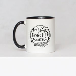 May include: White ceramic mug with a black handle and rim. The mug features the text "Mums make life Beautiful" in a black, decorative font, surrounded by a floral wreath design. The mug is a standard size for coffee or tea.