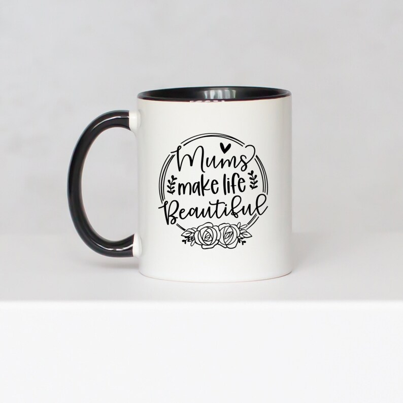 May include: White ceramic mug with a black handle and rim. The mug features the text "Mums make life Beautiful" in a black, decorative font, surrounded by a floral wreath design. The mug is a standard size for coffee or tea.