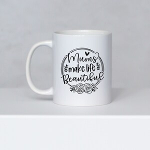 May include: White ceramic mug with a curved handle. The mug features the text "Mums make life Beautiful" in a black, decorative font, surrounded by a floral wreath design. The mug is set against a neutral background.