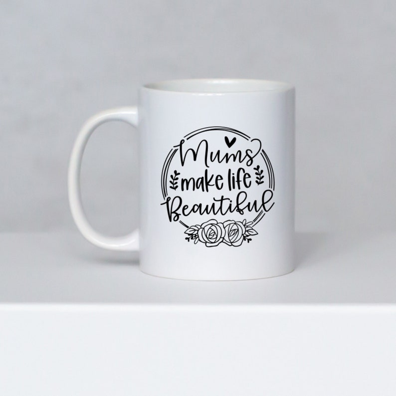 May include: White ceramic mug with a curved handle. The mug features the text "Mums make life Beautiful" in a black, decorative font, surrounded by a floral wreath design. The mug is set against a neutral background.