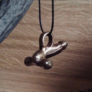 May include: A bronze pendant in the shape of a stylized figure with a ring at the top for attaching a necklace. The pendant is hanging from a black cord.