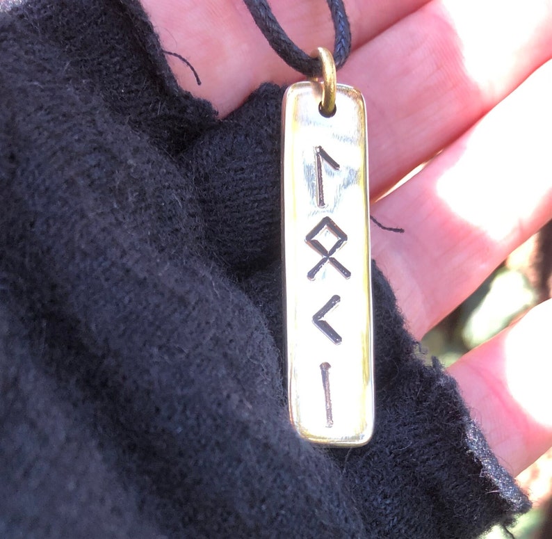 May include: A silver pendant with a black cord necklace. The pendant is rectangular and has four runes engraved on it.