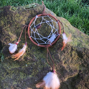 May include: A brown leather dreamcatcher with white string netting and brown feathers hanging from the bottom. The dreamcatcher is sitting on a mossy rock.