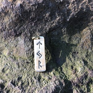 May include: A silver charm with four Norse runes engraved on it. The runes are:  "Algiz", "Sowulo", "Eihwaz", and "Raido".
