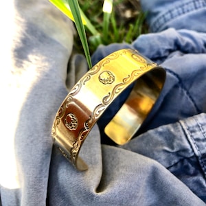 May include: A gold-toned metal bracelet with a skull design. The bracelet is engraved with a repeating pattern of skulls and swirls.