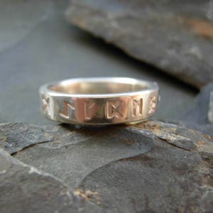 Op de afbeelding: Een zilveren ring met een band van gegraveerde runen. De ring ligt op een grijze steen.