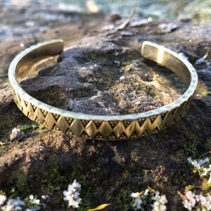 Peut inclure: Un bracelet de manchette en métal doré avec un motif géométrique. Le bracelet est sur une surface rocheuse texturée.