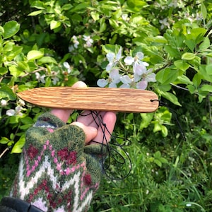 May include: A wooden knife with a dark grain pattern and a hole at the end, held by a person wearing a green and pink patterned glove. The knife is displayed against a backdrop of green foliage and white flowers.