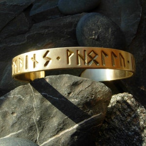 Puede incluir: Una pulsera de metal de color dorado con una inscripción en relieve de runas nórdicas. La inscripción dice "Hvíts - ÞNRMM - I".