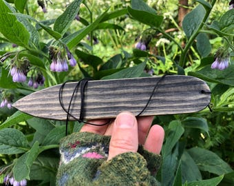 May include: A wooden, spear-shaped item with a black cord, held against a backdrop of green foliage and purple flowers. The wood has a gray and black striped pattern. The item appears to be handmade.