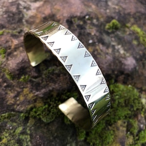 May include: A gold-toned metal bracelet with a geometric pattern of triangles. The bracelet is open and has a smooth, polished finish.