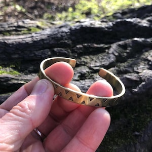 Könnte beinhalten: Ein goldfarbenes Armreif-Armband mit einer Reihe kleiner, dunkler Dreieckmuster. Das offene Armband wird in einer Hand gehalten und zeigt das geometrische Design. Das Armband ist aus Metall.