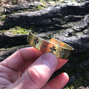 Op de afbeelding: Een goudkleurige manchetarmband met geëtste geometrische patronen. Het open manchetontwerp heeft een reeks herhalende vierkante en ruitvormige vormen. De armband wordt in een hand gehouden en toont zijn gepolijste afwerking en ingewikkelde details.