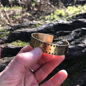 Op de afbeelding: Een brede, goudkleurige manchetarmband met een geometrisch patroon van driehoeken en stippen. De armband wordt in een hand gehouden, waarbij het open ontwerp wordt getoond. De sieraden zijn van metaal en hebben een vintage uitstraling.