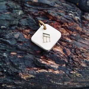 May include: A silver square charm with a small, engraved symbol. The charm is hanging from a gold loop.