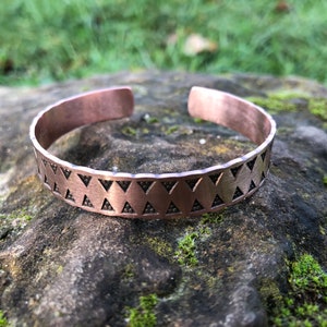 May include: A copper bracelet with a geometric pattern of triangles. The bracelet is on a rock with moss.