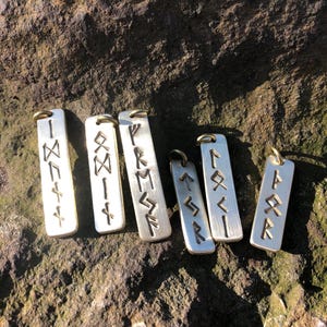 May include: Six silver metal pendants with engraved runes. The pendants are rectangular and have a hole at the top for hanging.