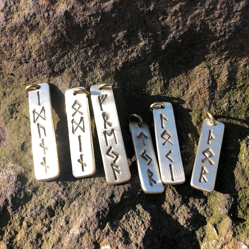 May include: Six silver metal pendants with engraved runes. The pendants are rectangular and have a hole at the top for hanging.