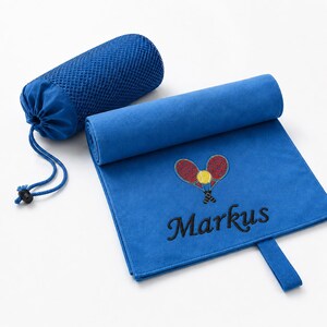 Op de afbeelding: Een koningsblauwe microvezel handdoek met een geborduurd ontwerp van twee gekruiste tennisrackets en een gele tennisbal, met de naam Markus in zwarte draad eronder gestikt. De handdoek bevat een bijpassende blauwe mesh tas met trekkoord.