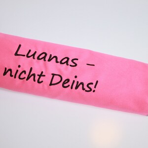 Op de afbeelding: Een felroze cilindervormige stoffen etui met de zwart geborduurde tekst "Luanas &ndash; nicht Deins!". De etui is voorzien van een grijze gestikte rand en een kleine grijze lus aan &eacute;&eacute;n uiteinde, die dient als een functioneel sluitingsmechanisme.