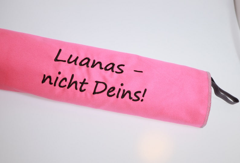 Op de afbeelding: Een felroze cilindervormige stoffen etui met de zwart geborduurde tekst "Luanas &ndash; nicht Deins!". De etui is voorzien van een grijze gestikte rand en een kleine grijze lus aan &eacute;&eacute;n uiteinde, die dient als een functioneel sluitingsmechanisme.