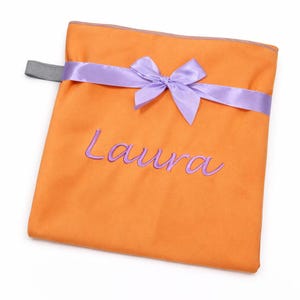 Op de afbeelding: Een vierkant oranje stoffen zakje met een lavendelkleurig satijnen lint dat aan de bovenkant tot een strik is geknoopt. De naam Laura is in lavendelkleurig garen op de voorzijde geborduurd. Een kleine grijze elastische lus zit aan de linkerkant.