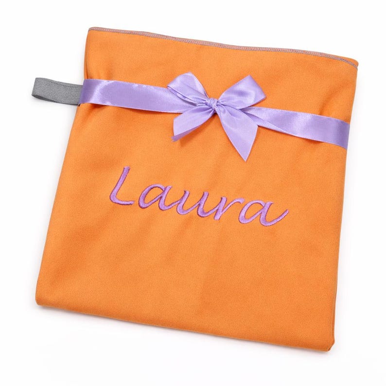 Op de afbeelding: Een vierkant oranje stoffen zakje met een lavendelkleurig satijnen lint dat aan de bovenkant tot een strik is geknoopt. De naam Laura is in lavendelkleurig garen op de voorzijde geborduurd. Een kleine grijze elastische lus zit aan de linkerkant.