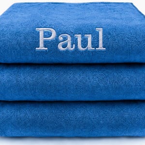 Op de afbeelding: Een stapel van drie gevouwen koningsblauwe handdoeken. De bovenste handdoek is voorzien van de naam Paul, geborduurd in witte letters met schreef in hoofdletters en kleine letters, gecentreerd op de voorzijde. De handdoeken hebben een textuur.