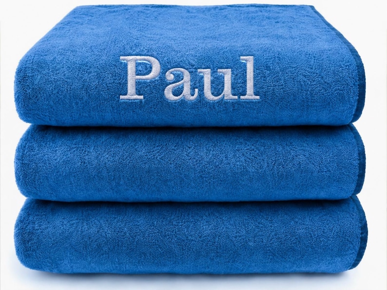 Op de afbeelding: Een stapel van drie gevouwen koningsblauwe handdoeken. De bovenste handdoek is voorzien van de naam Paul, geborduurd in witte letters met schreef in hoofdletters en kleine letters, gecentreerd op de voorzijde. De handdoeken hebben een textuur.