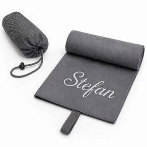 Op de afbeelding: Een donkergrijze microvezel handdoek met de geborduurde naam Stefan in witte schrijfletters. De handdoek is gedeeltelijk opgerold en bevat een bijpassend donkergrijs mesh opbergzakje met trekkoord en een zwarte plastic koordstopper.