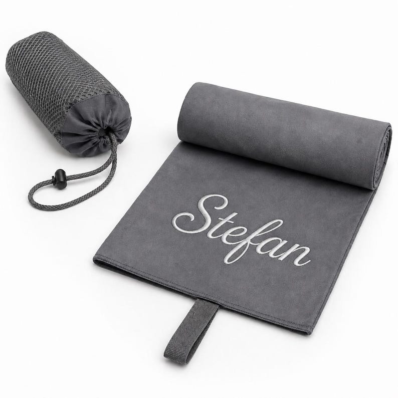 Op de afbeelding: Een donkergrijze microvezel handdoek met de geborduurde naam Stefan in witte schrijfletters. De handdoek is gedeeltelijk opgerold en bevat een bijpassend donkergrijs mesh opbergzakje met trekkoord en een zwarte plastic koordstopper.