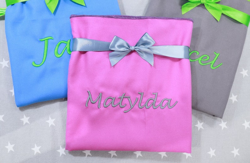 Op de afbeelding: Drie gevouwen stoffen zakjes in blauw, roze en grijs. Het roze zakje is voorzien van de naam Matylda geborduurd in grijze draad en is gestrikt met een zilveren satijnen lint. De blauwe en grijze zakjes hebben groene linten en borduursels.