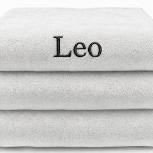 Op de afbeelding: Een stapel van drie gevouwen witte handdoeken met een zachte, pluche textuur. De bovenste handdoek is voorzien van de naam Leo, geborduurd in zwarte schreefletters in het midden van de voorzijde. De handdoeken zijn netjes op elkaar gestapeld.
