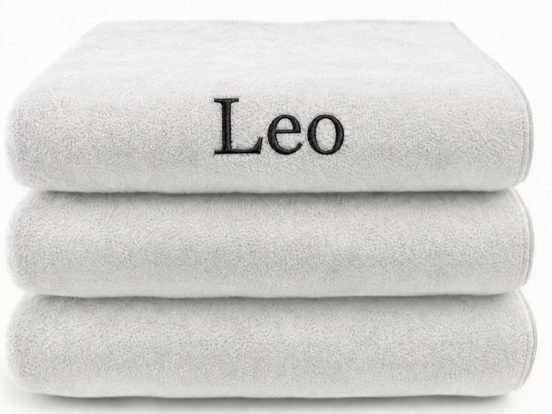 Op de afbeelding: Een stapel van drie gevouwen witte handdoeken met een zachte, pluche textuur. De bovenste handdoek is voorzien van de naam Leo, geborduurd in zwarte schreefletters in het midden van de voorzijde. De handdoeken zijn netjes op elkaar gestapeld.