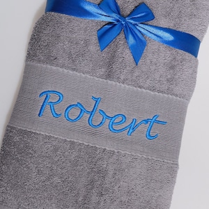 Peut inclure: Une serviette de bain grise avec un ruban bleu noué autour. La serviette est brodée du nom "Robert" en fil bleu.