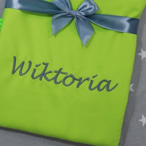 Op de afbeelding: Een fel limoengroen stoffen item met een donkergrijze satijnen strik aan de bovenkant. De naam Wiktoria is in donkergrijs cursief schrift geborduurd in het midden van het limoengroene oppervlak, wat zorgt voor een duidelijk contrast.