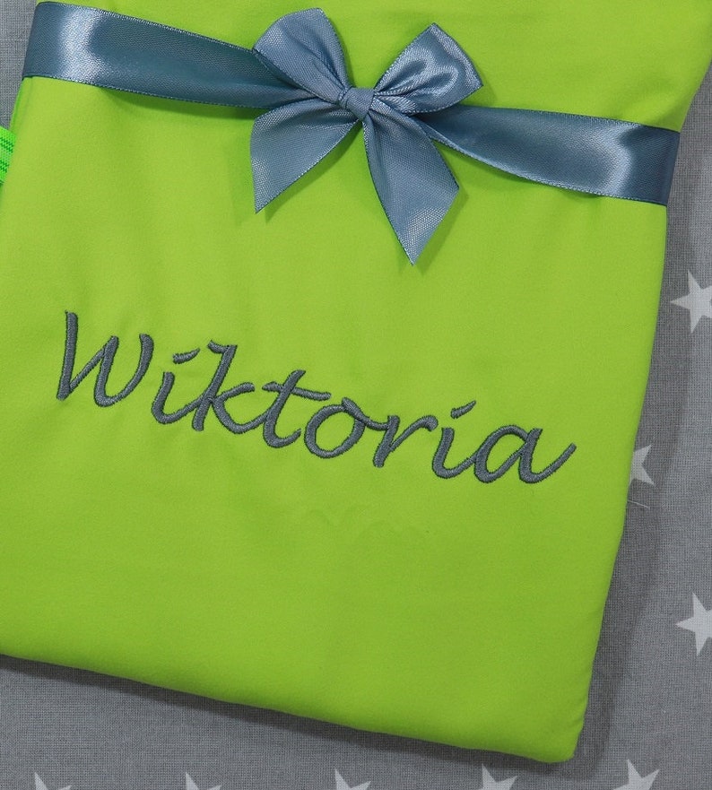 Op de afbeelding: Een fel limoengroen stoffen item met een donkergrijze satijnen strik aan de bovenkant. De naam Wiktoria is in donkergrijs cursief schrift geborduurd in het midden van het limoengroene oppervlak, wat zorgt voor een duidelijk contrast.