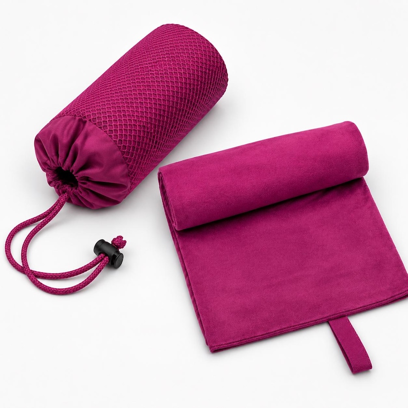 Op de afbeelding: Een magenta microvezel handdoek gevouwen in een rechthoekige vorm met een klein stoffen lusje aan &eacute;&eacute;n rand, vergezeld van een bijpassend cilindervormig magenta mesh opbergzakje met een trekkoordsluiting en een zwarte plastic stopper.
