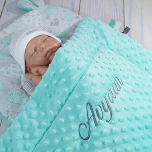 Peut inclure: Une couverture de bébé turquoise avec un motif de cœurs blancs et gris. La couverture est brodée du nom "Aayaan".