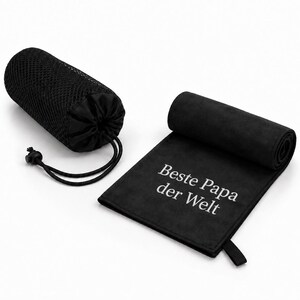 Op de afbeelding: Een zwarte microvezel handdoek met de witte tekst "Beste Papa der Welt" erop gedrukt, gedeeltelijk opgerold, vergezeld van een zwarte mesh opbergtas met trekkoordsluiting, ontworpen voor compacte draagbaarheid en opslag.